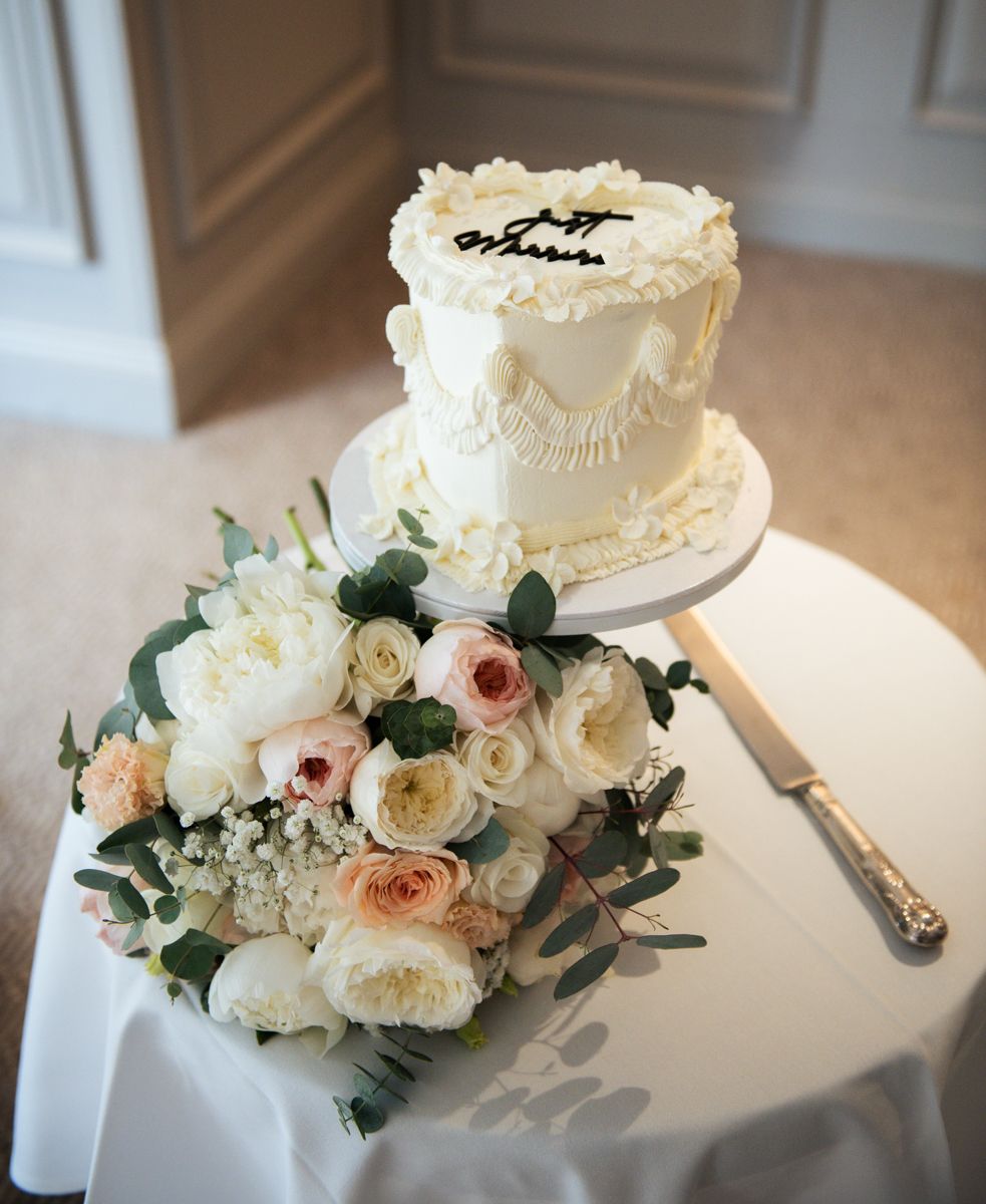 Two-tier white wedding cake with Just Married topper beside a bouquet of white and blush roses on an elegant table.