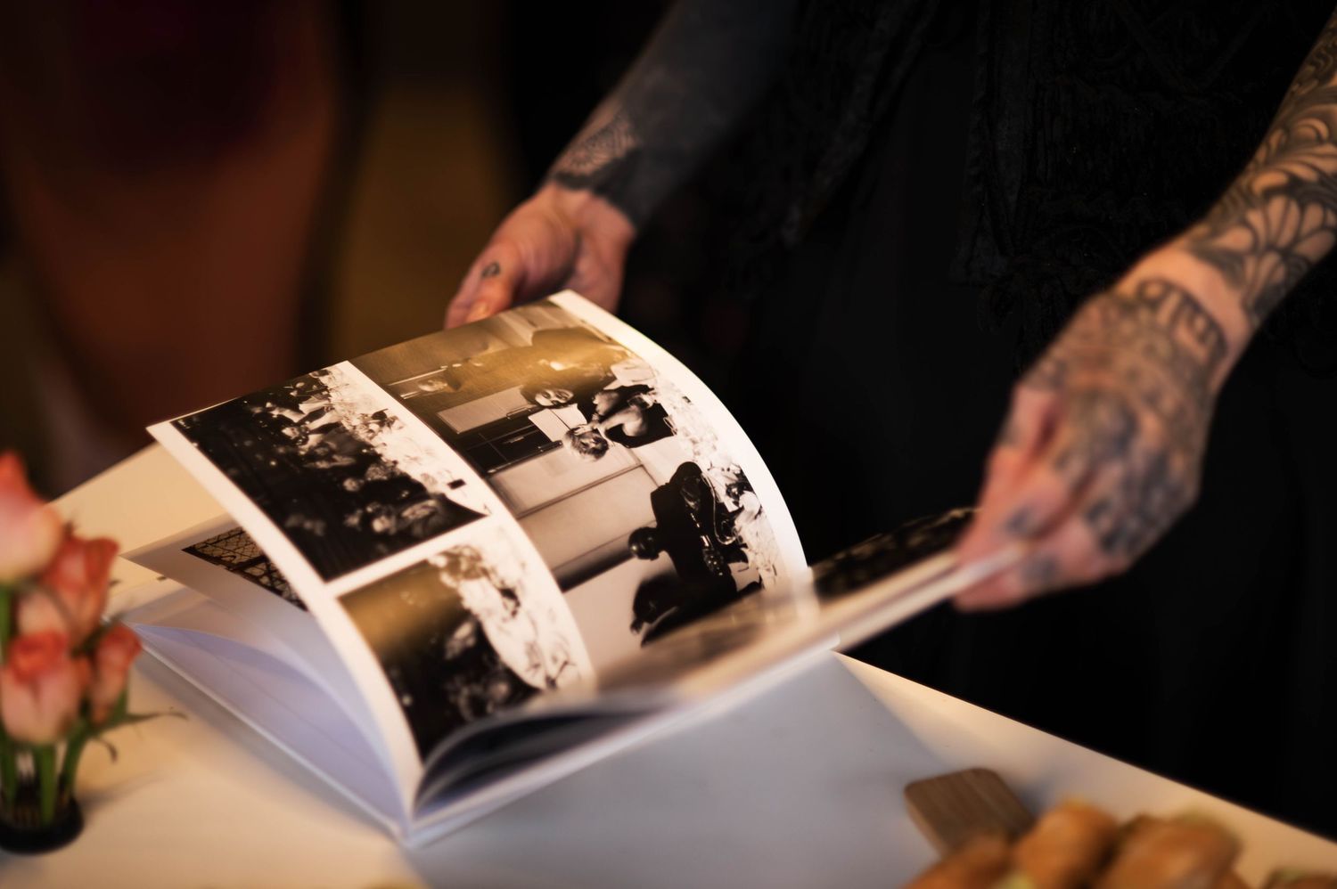 Tattooed hands flip through a black and white photo album on a white table with pink flowers in the background.