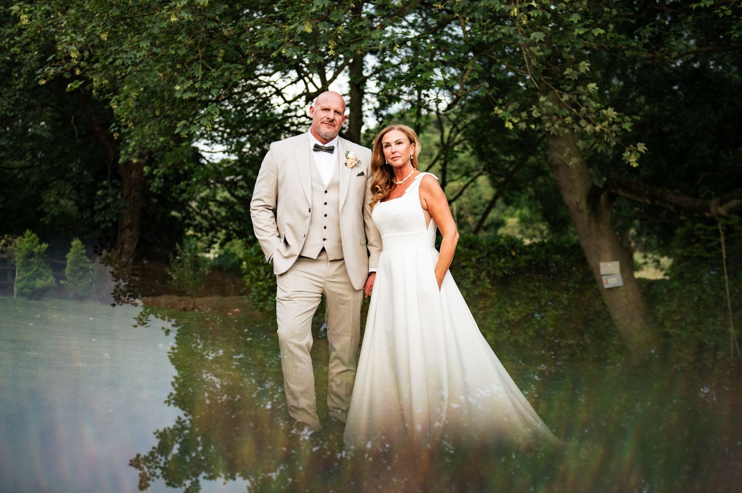 Bride in white gown and groom in tan suit pose together outdoors near a pond surrounded by lush green trees.