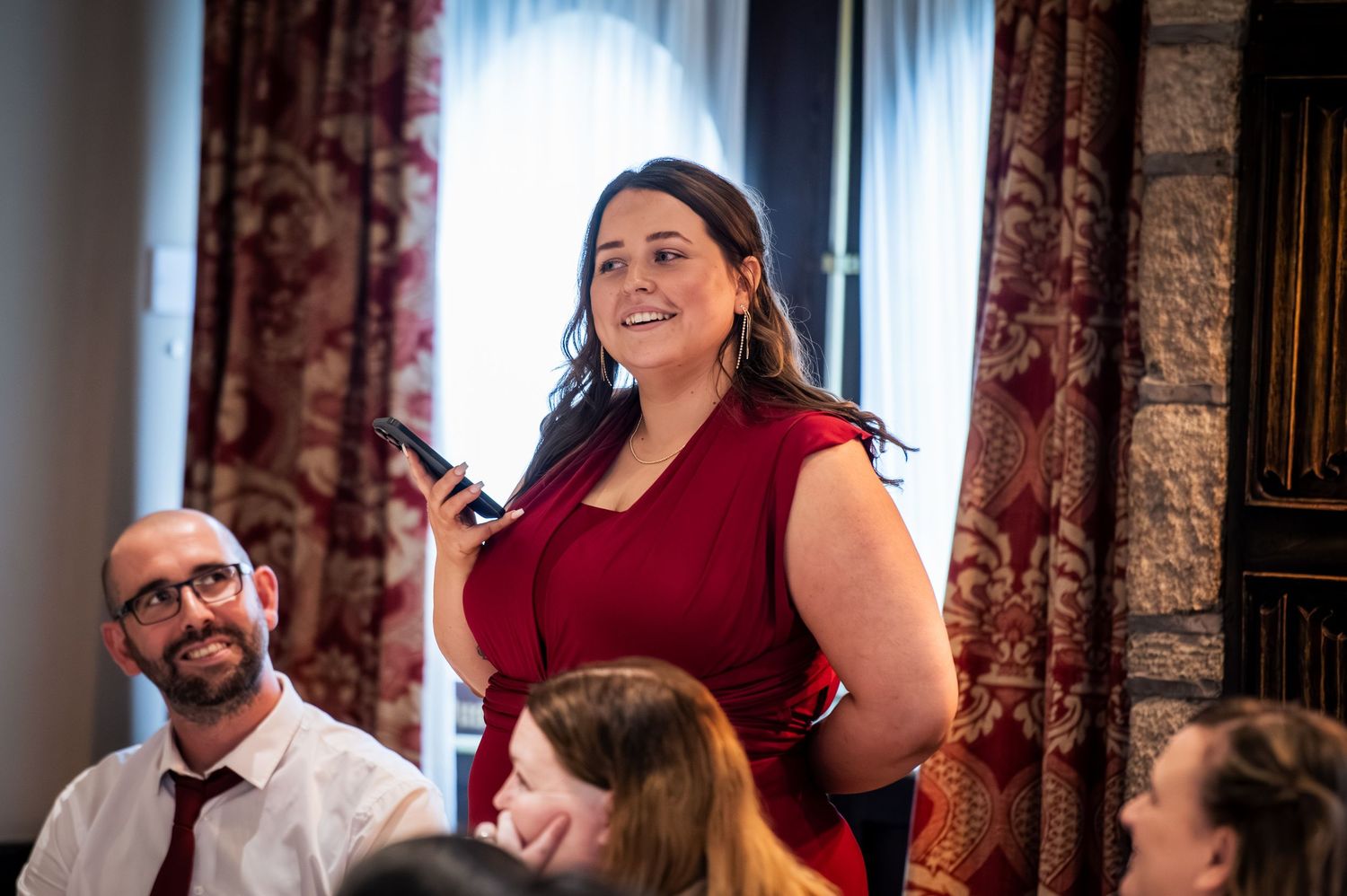 Woman in red dress speaking at formal event, smiling confidently while seated guests listen attentively in elegant venue.