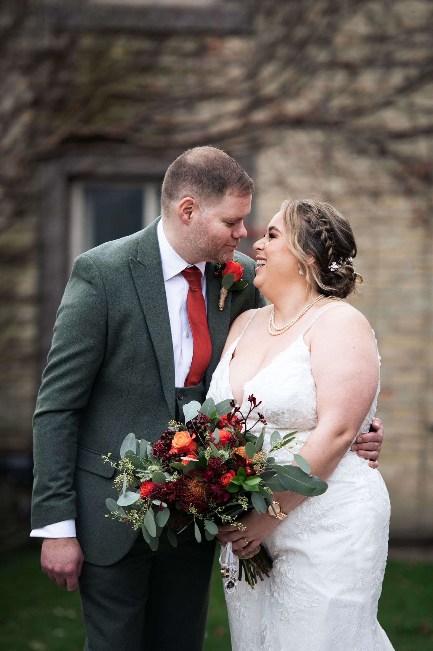 Romantic wedding couple with red bouquet poses against stone wall backdrop at the Fleece Inn, Halifax.