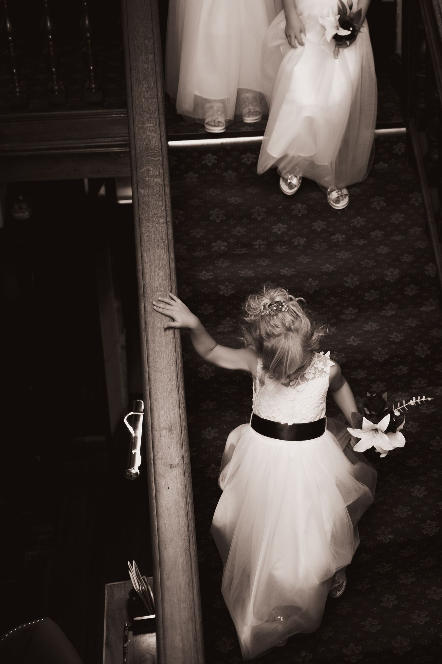 Young flower girl in white dress with black sash holds flowers at a wedding ceremony, sepia tone.