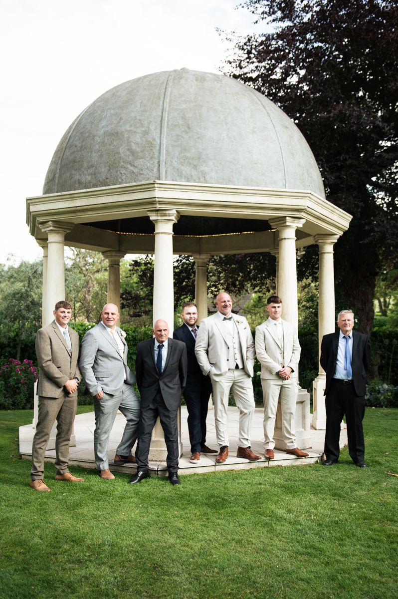 Seven men in suits posing together under a domed gazebo in a lush garden setting.