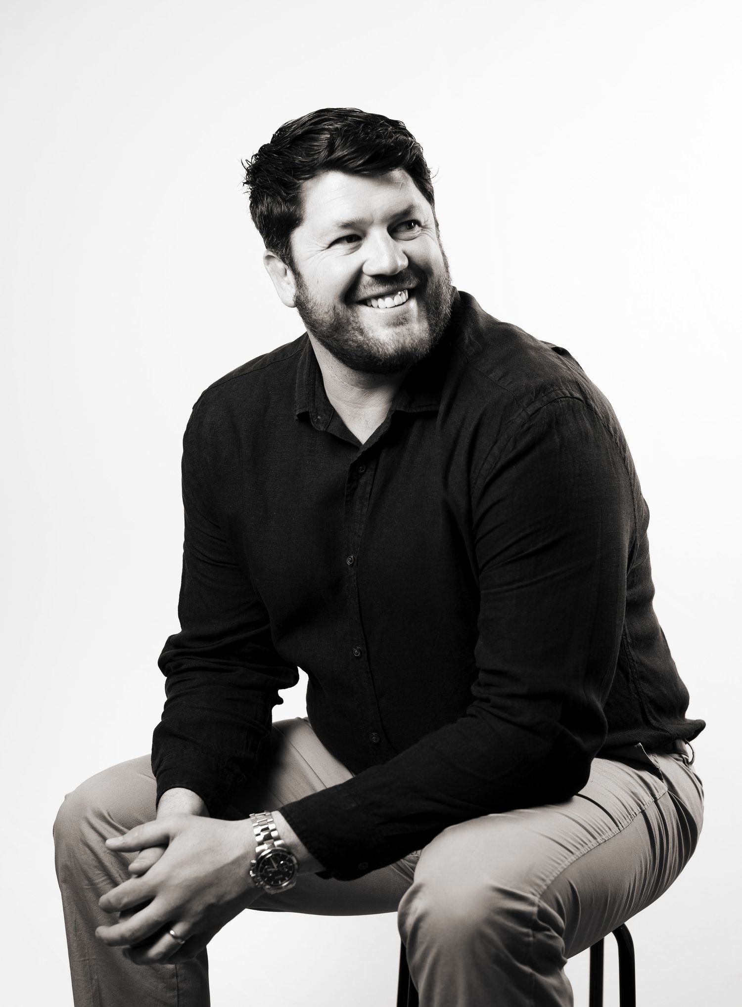 Business headshot of a black and white portrait of a man in dark clothing sitting on a stool, taken in Leeds.