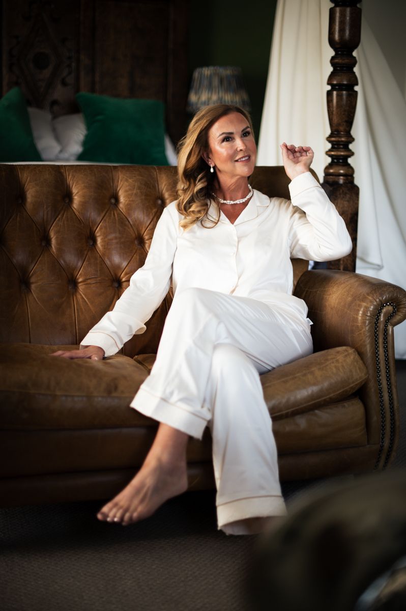 Woman in white silk pajamas relaxing on brown leather tufted sofa, smiling with pearl necklace and wavy blonde hair.
