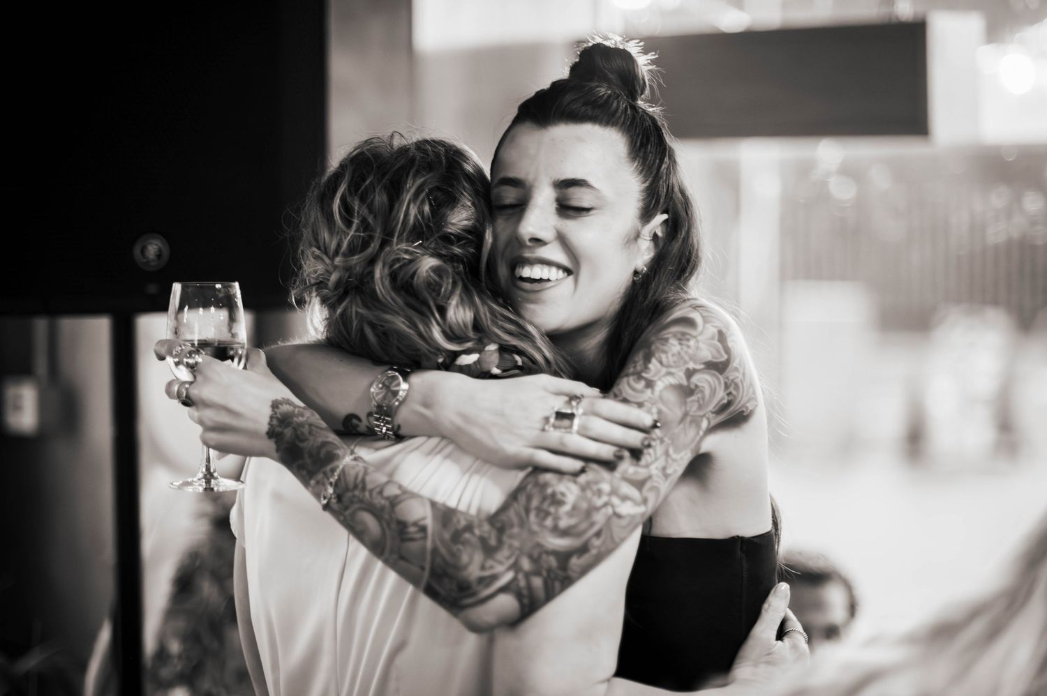 Two women with tattoos share a joyful hug at a wedding reception, one holding a wine glass, in black and white.