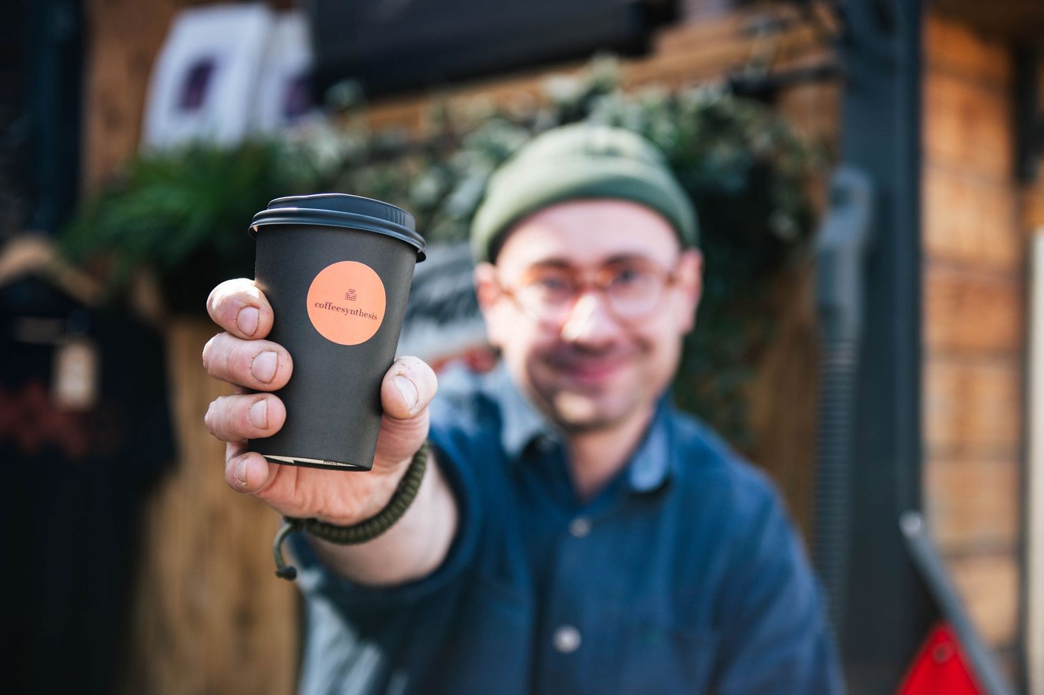 A person holds up a black coffee cup with an orange logo outside a wooden storefront for business photos in Leeds.