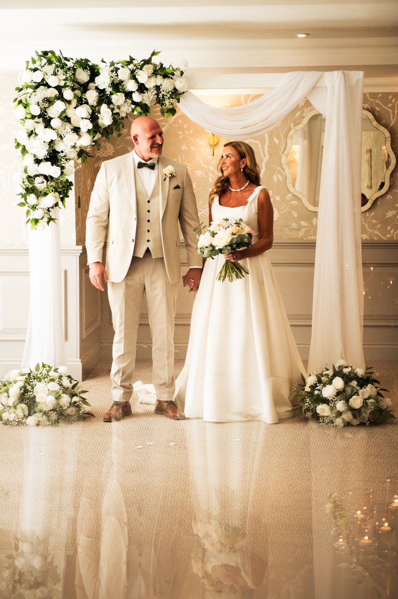 Elegant wedding couple standing under floral arch with white roses, bride in gown holding bouquet, groom in cream suit.