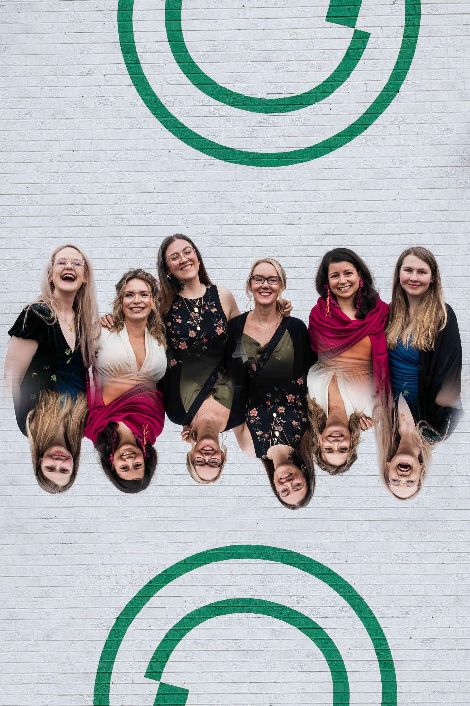 Group of seven smiling women posing together against a white brick wall with green circle designs.