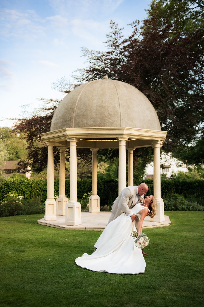 Bride and groom share a romantic dip kiss in front of an elegant stone gazebo surrounded by lush gardens.