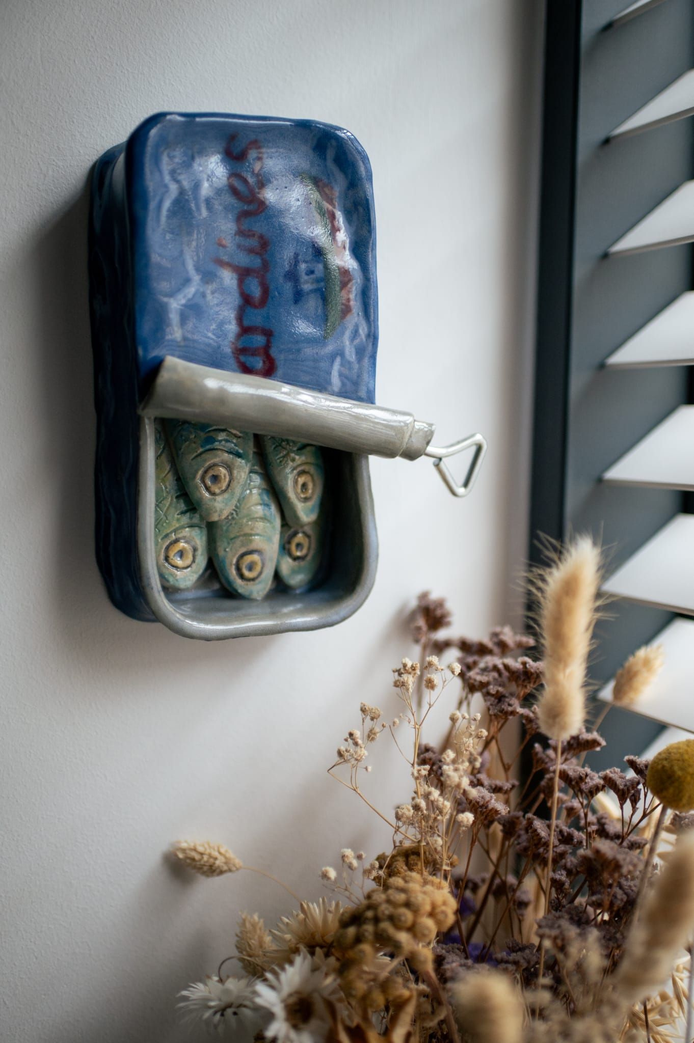 Decorative dried flowers beneath a pottery sardine tin mounted on a white wall with window blinds.