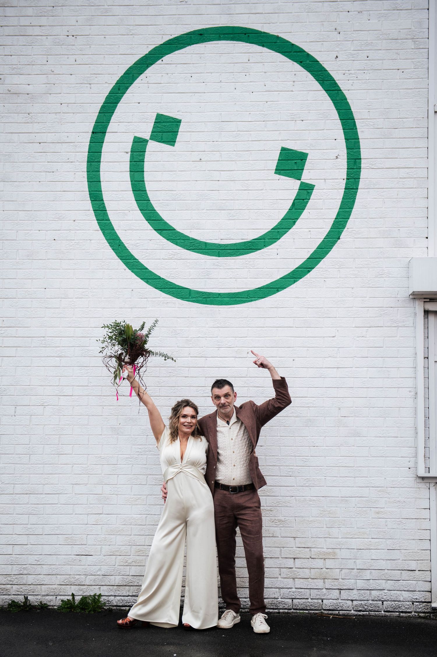 Bride and groom celebrating in front of a green smiley face mural on a white brick wall.