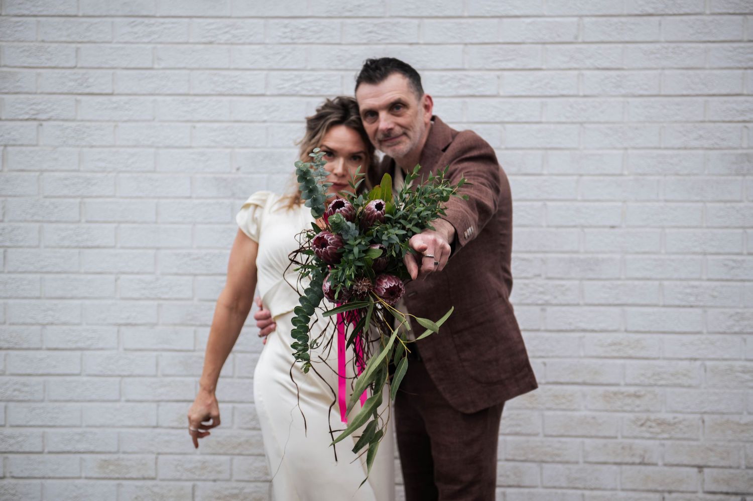 Bride and groom posing against white brick wall, groom extending lush green and red bridal bouquet with pink ribbon toward camera.