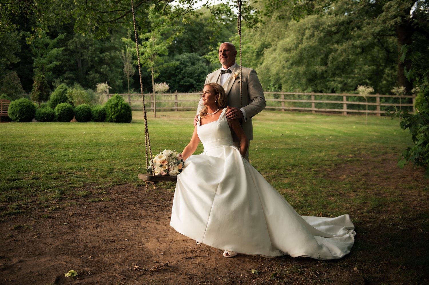 Bride in white gown and groom in gray suit pose near a tree swing in a lush green garden setting.