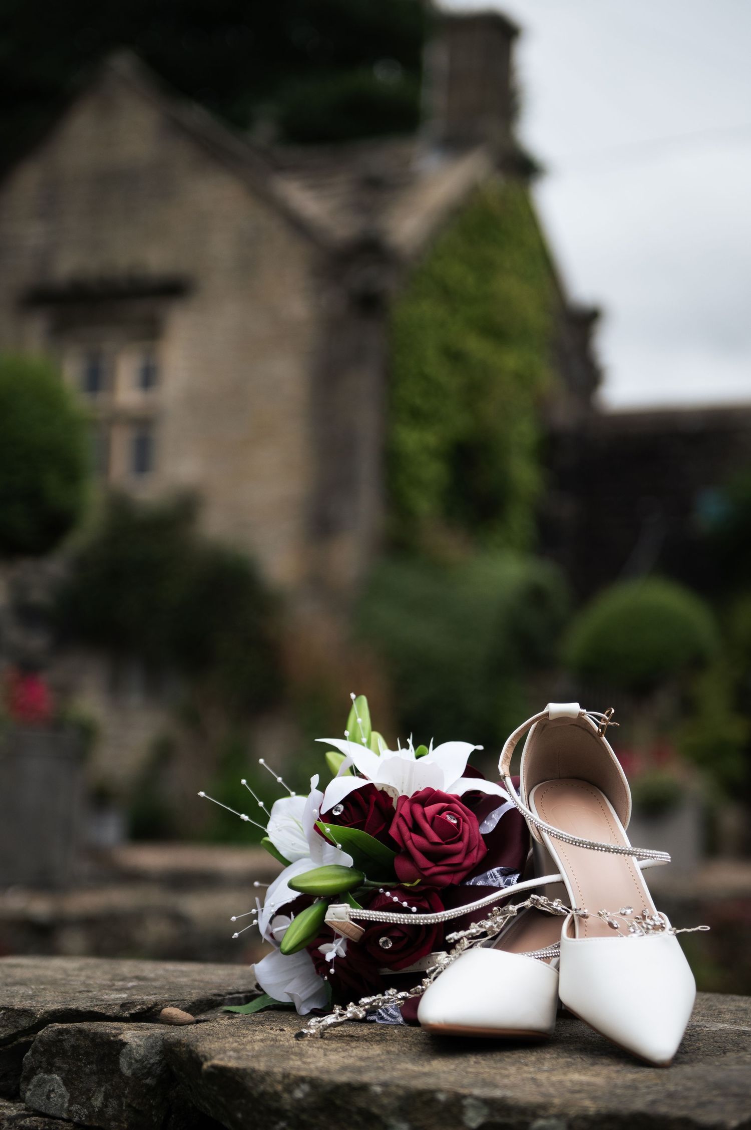 White bridal heels and a bouquet of red and white roses on stone steps with a blurred historic building backdrop.