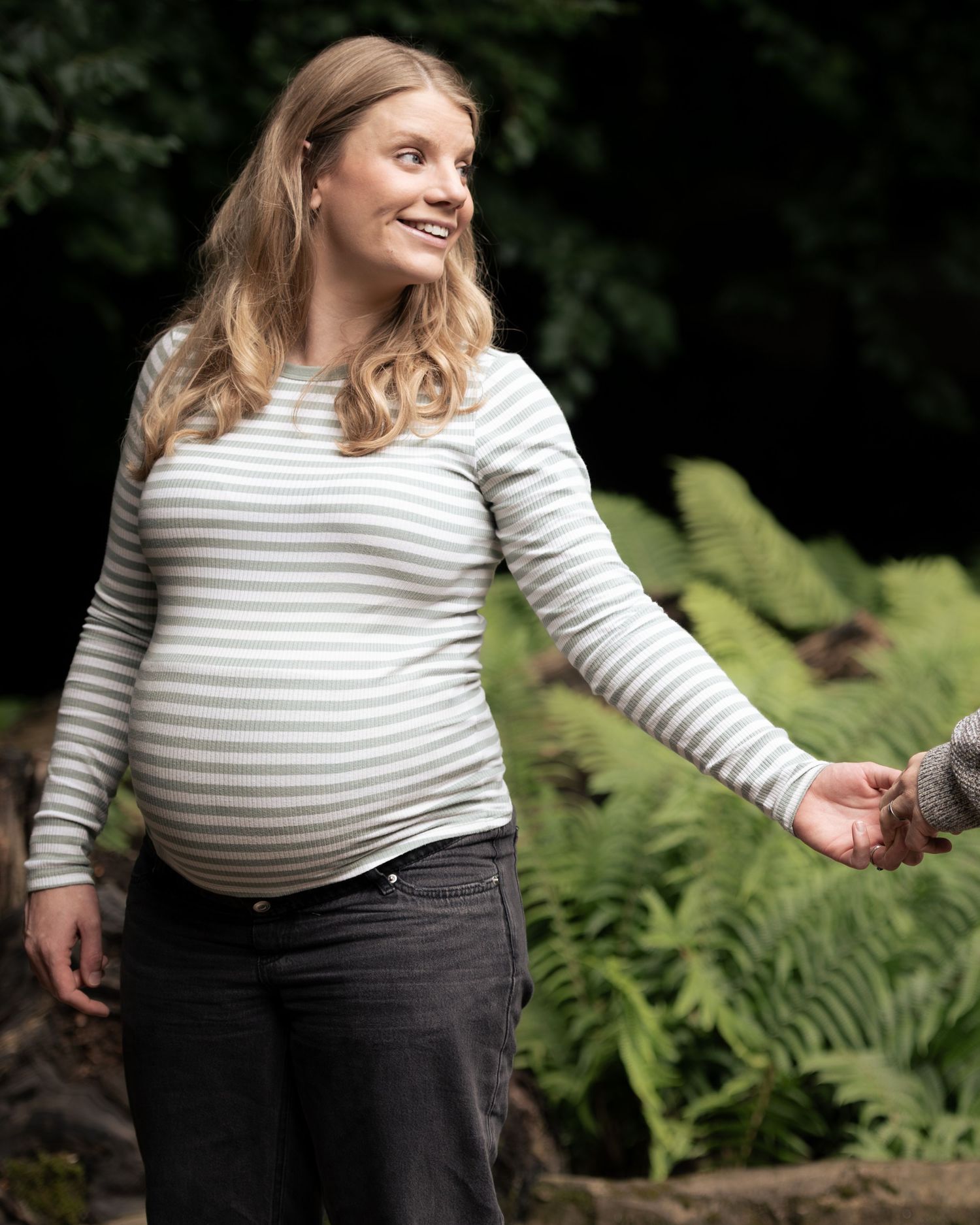 A mother in a striped shirt smiles warmly while posing against a natural backdrop with ferns, during a maternity shoot in Leeds.