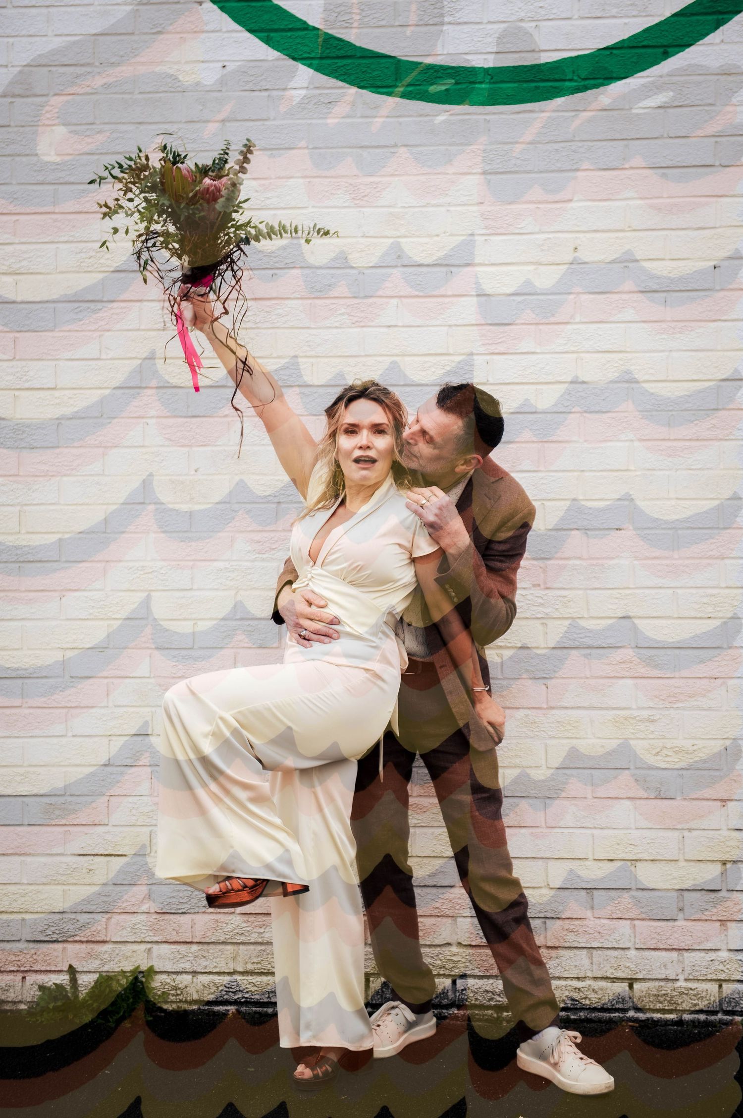 Joyful bride in white jumpsuit holds bouquet aloft while groom embraces her before a colorful mural wall.