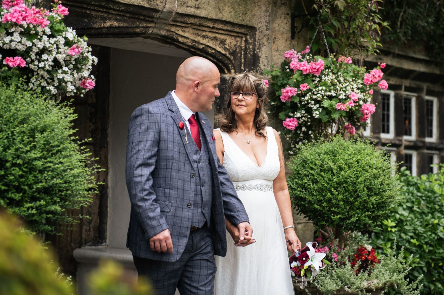 Bride and groom holding hands outside a stone venue adorned with pink and red flowers on their wedding day.