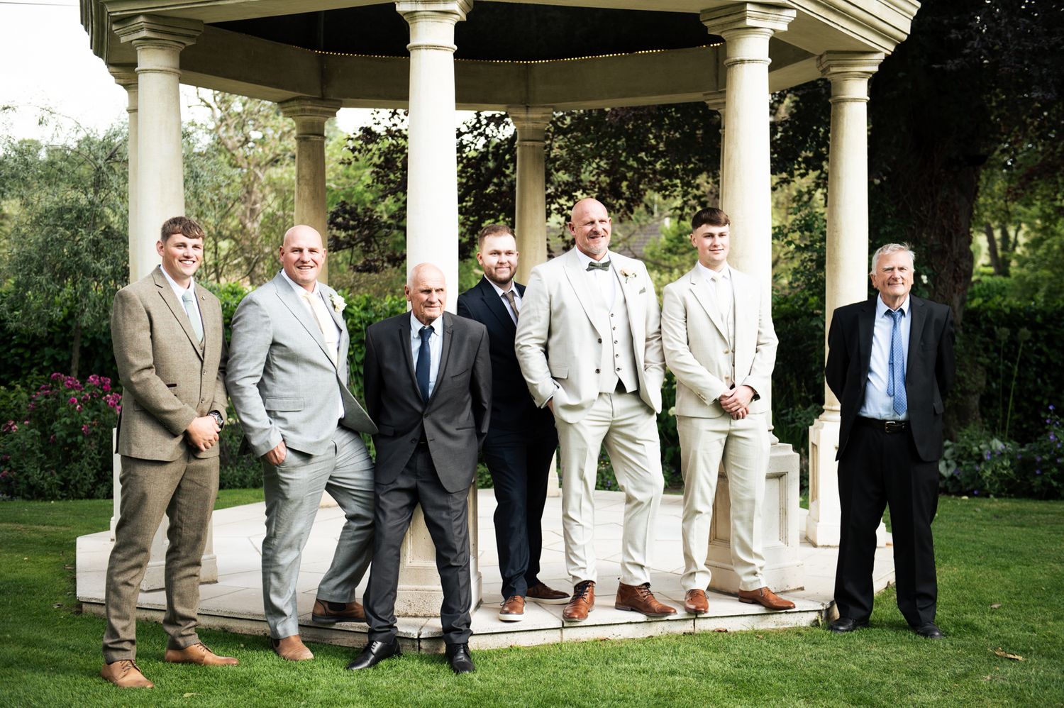 Group of seven men in suits posing together under a white gazebo in a lush garden setting.