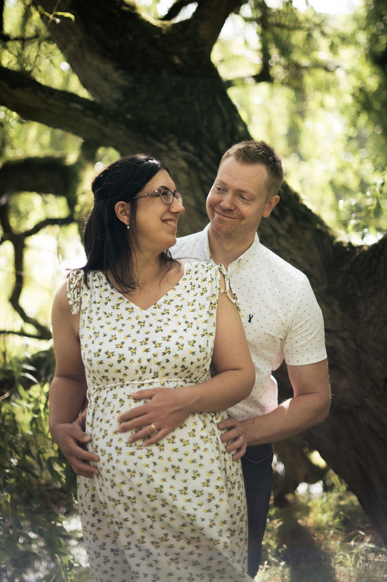 A couple smiles at one another under a willow tree during an outdoor maternity shoot in Leeds.