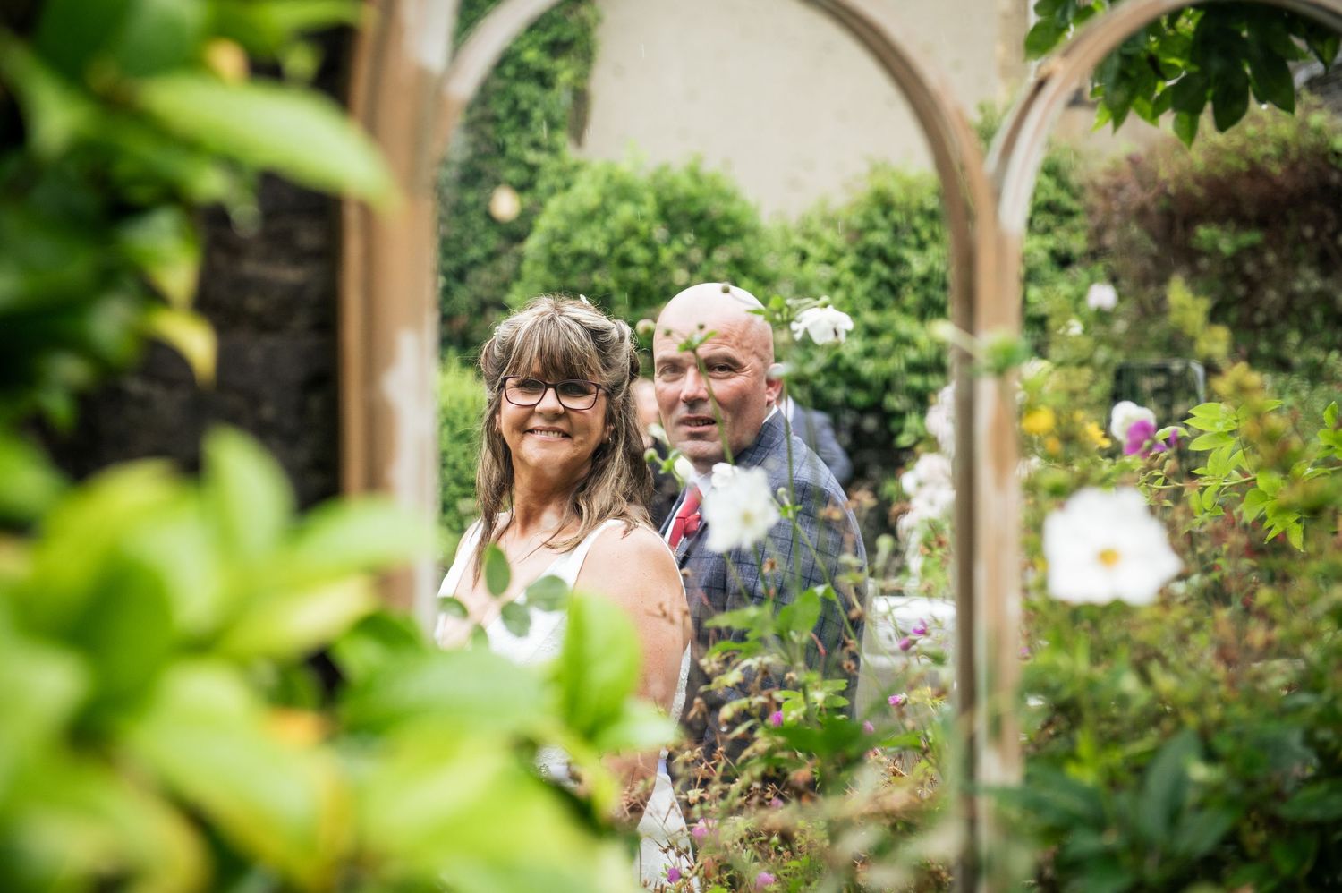 Couple smiling in a lush garden framed by a wooden arch with roses and greenery.