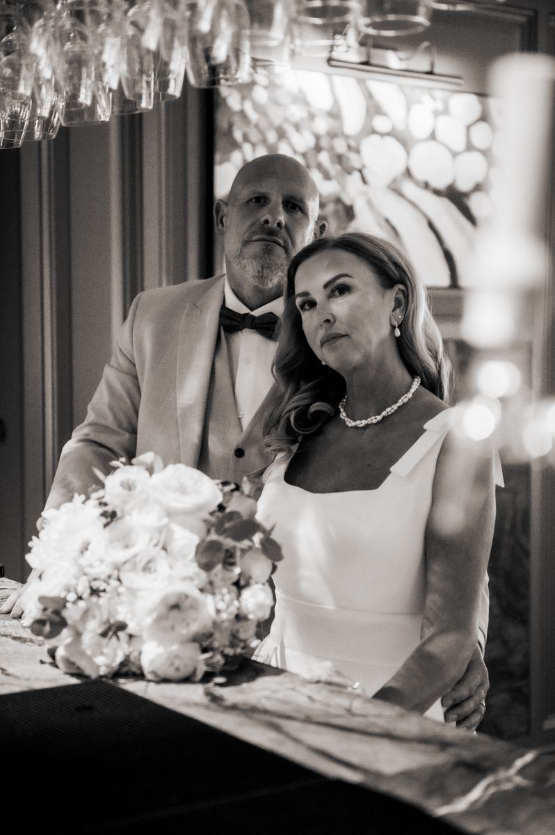 Black and white wedding photo of bride in white gown with bouquet and groom in tuxedo at elegant venue.