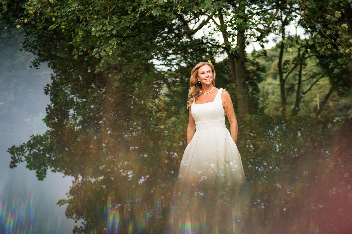 A bride in a white gown stands elegantly among lush green trees with a dreamy rainbow lens flare effect.