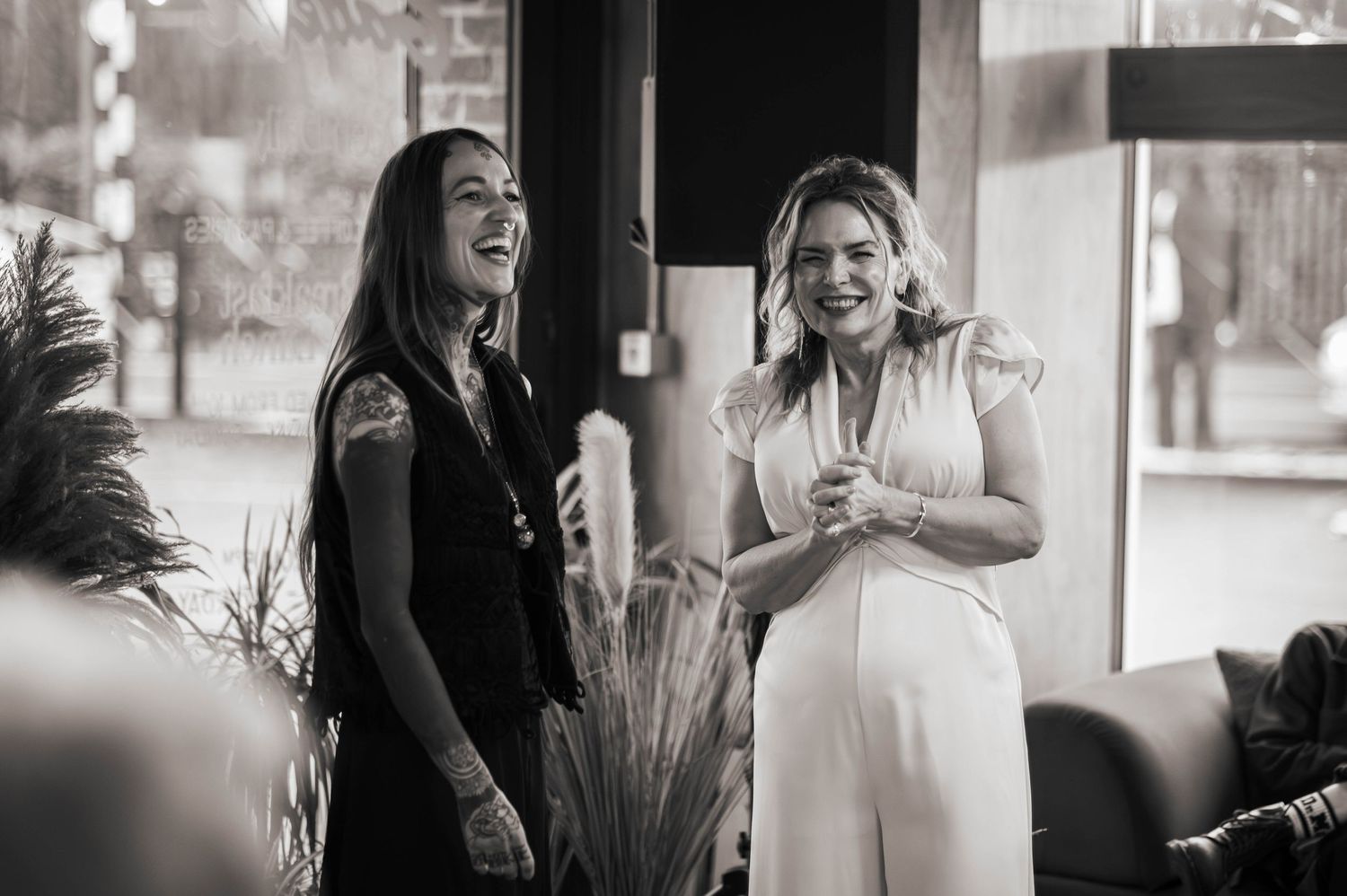 Two women laughing and smiling together at an indoor social gathering, captured in black and white photography.