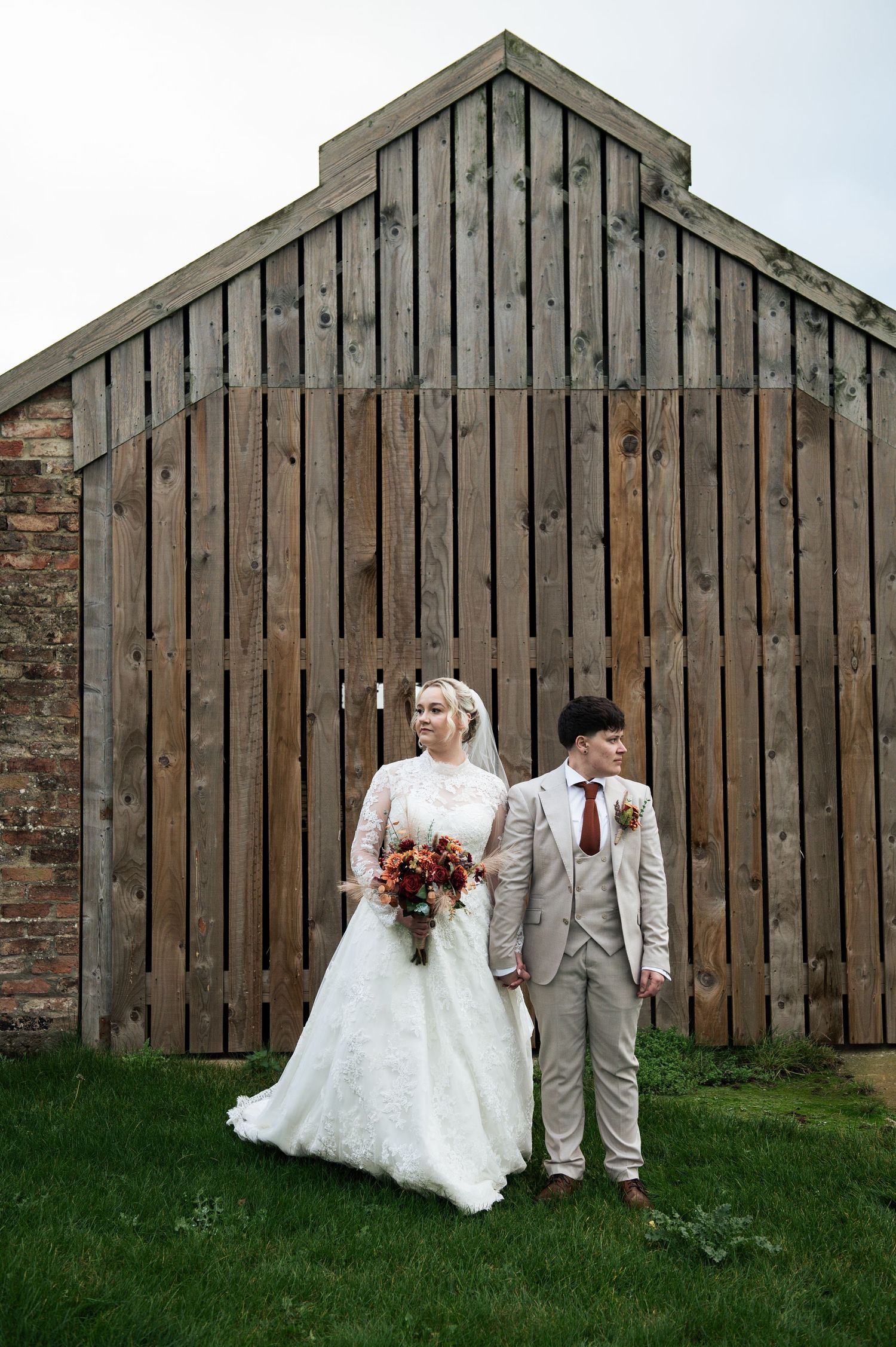 An LGBT wedding couple poses in front of a rustic wooden barn wall during their countryside celebration at Lodge Farm Weddings, Yorkshire.