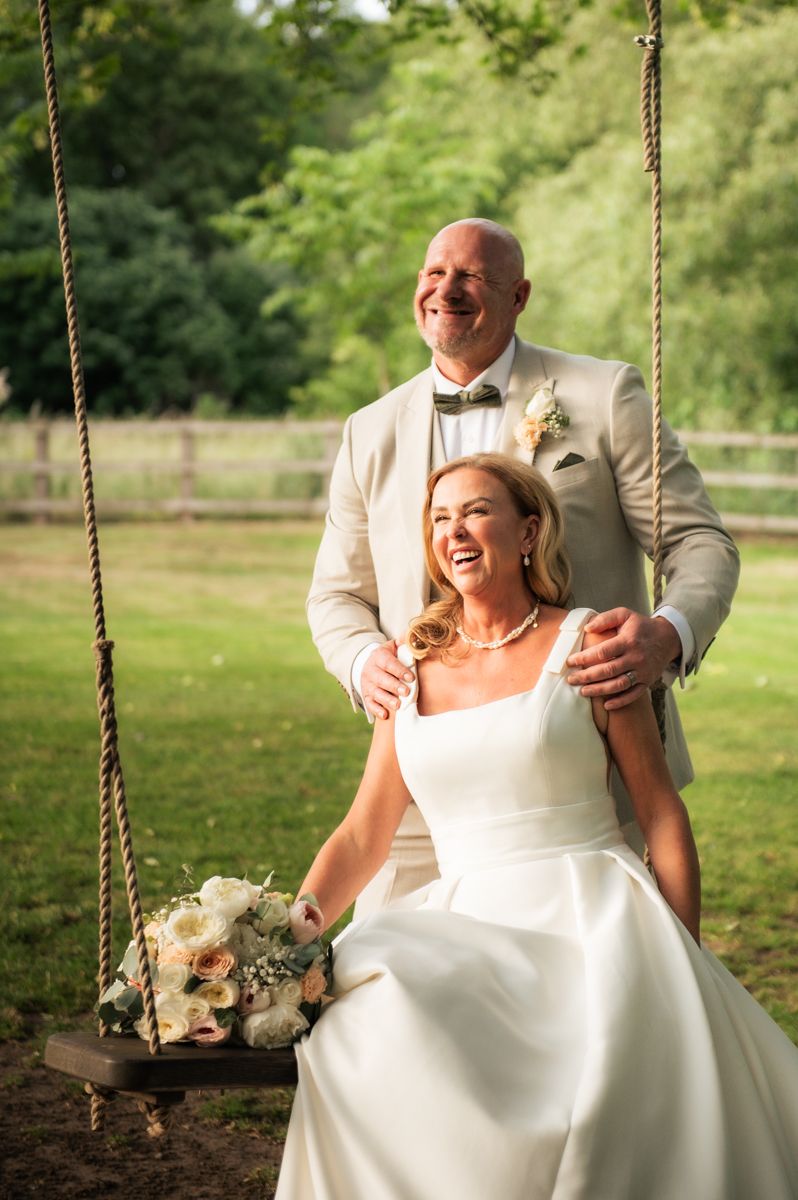 Bride sitting on a rope swing holding a bouquet while groom stands behind her, both laughing outdoors.