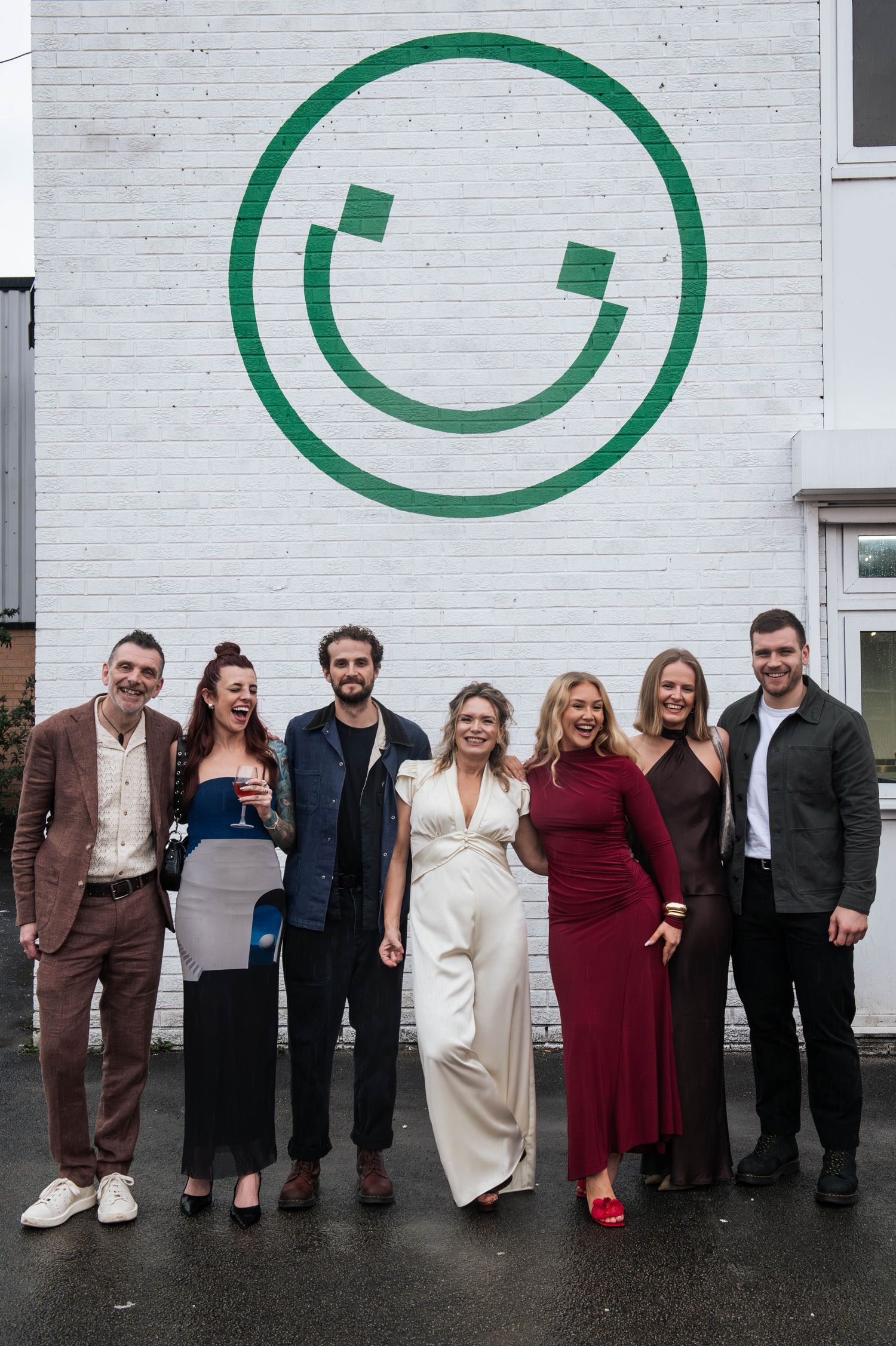 Group of seven people posing and smiling in front of a white brick wall with a green smiley face mural.