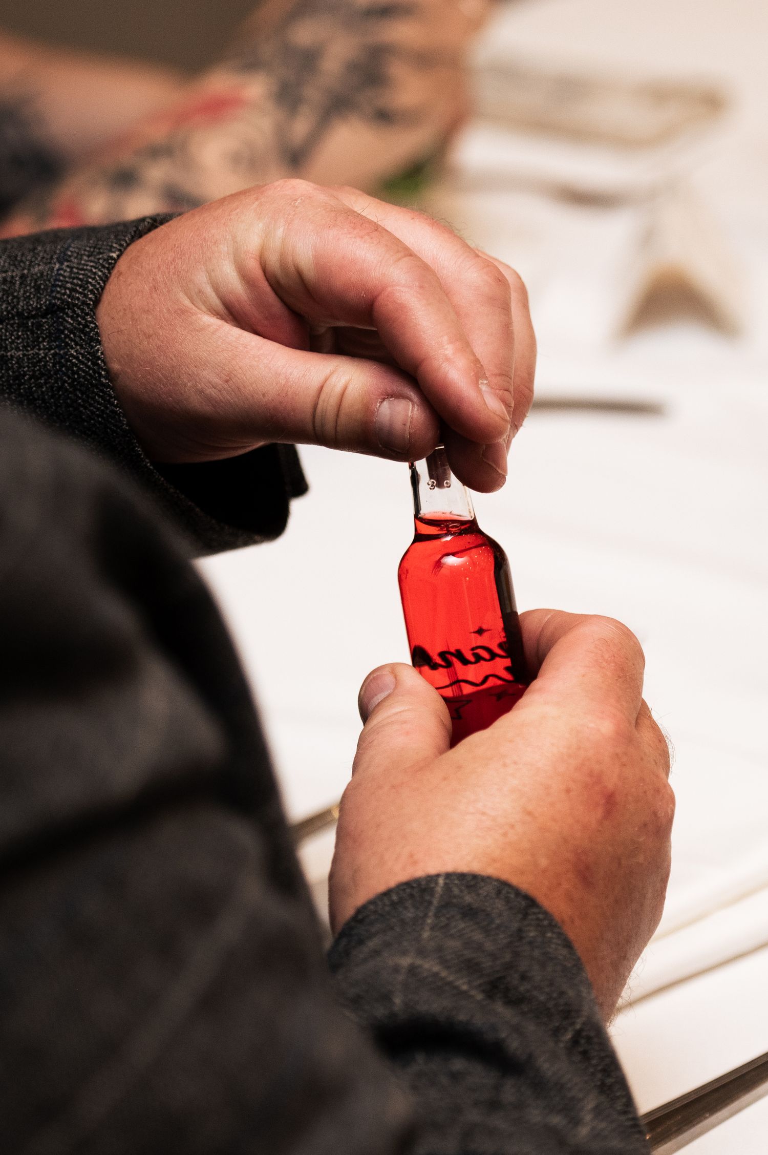 Hands opening a small red glass bottle with a dropper cap on a white surface.