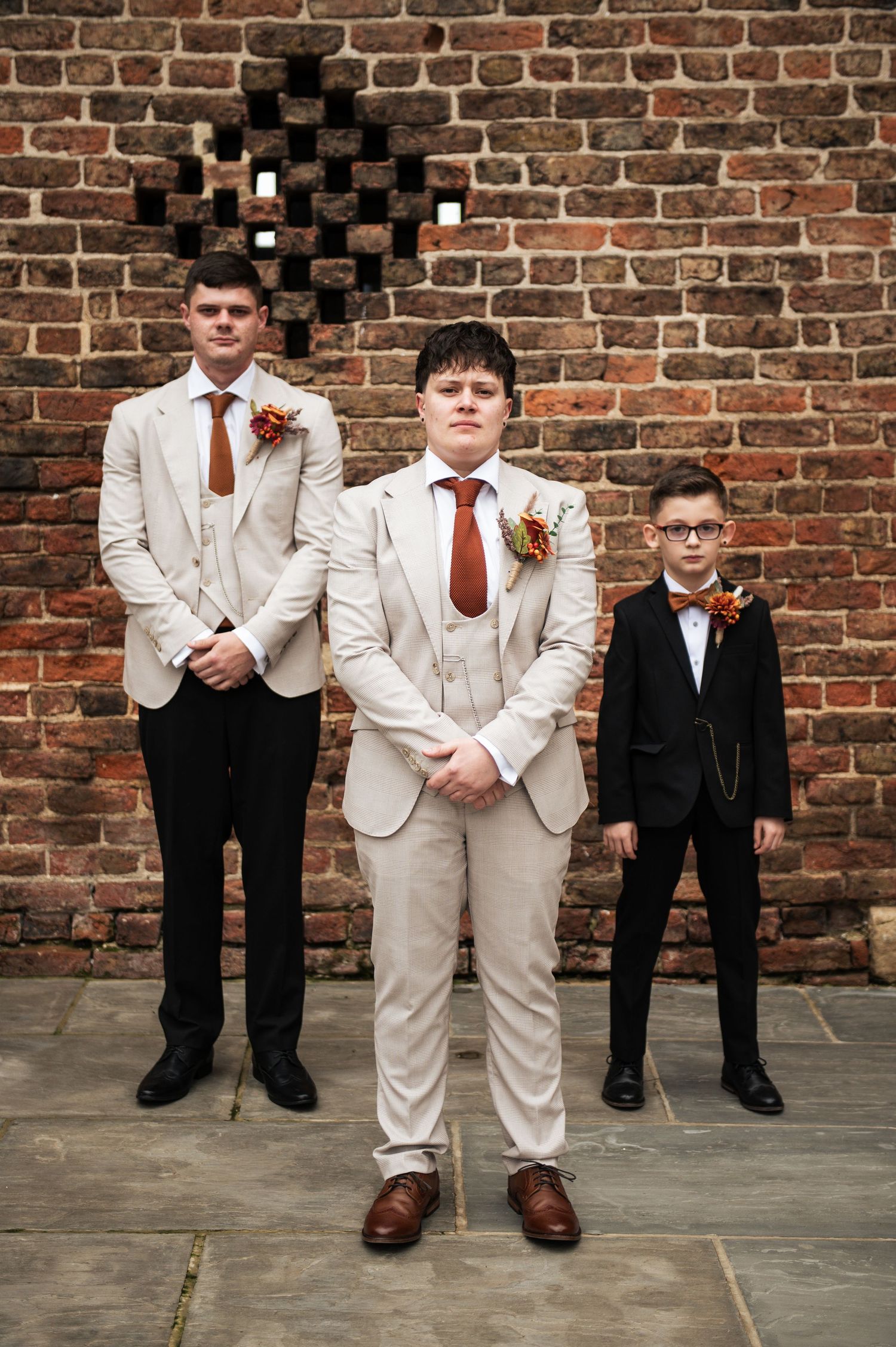 Groomsmen in beige suits and red ties pose formally against a rustic brick wall, at Lodge Farm Weddings, Yorkshire.