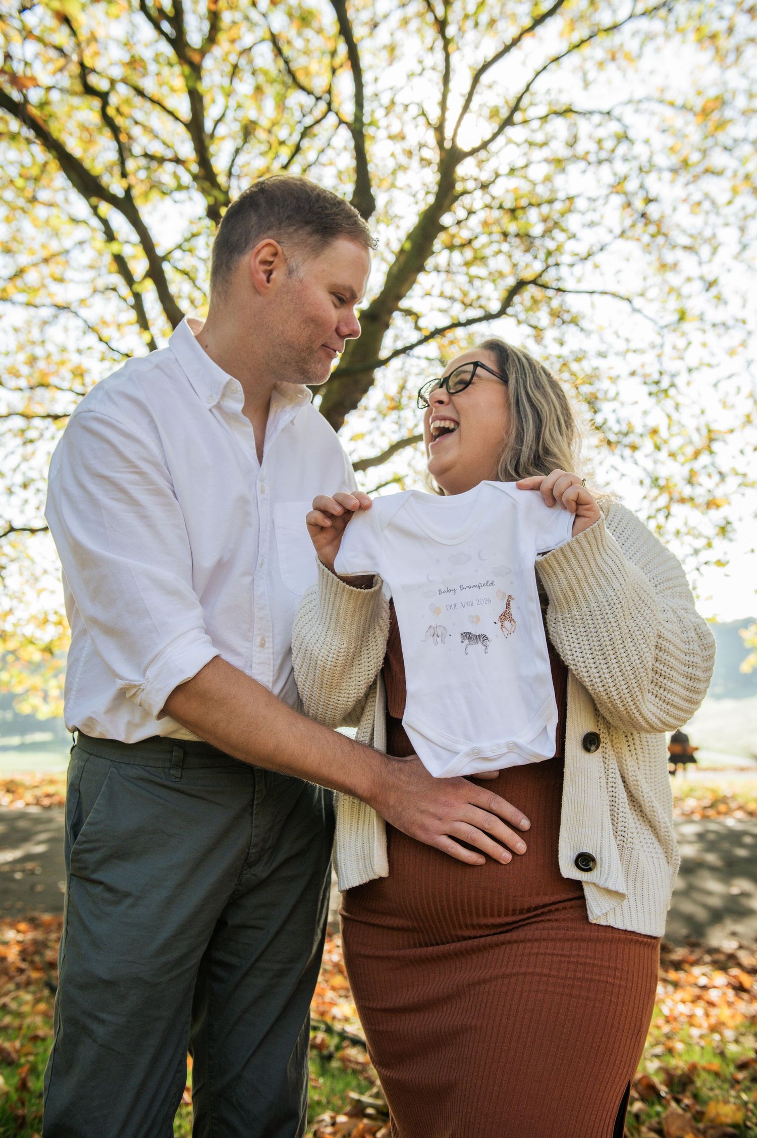 A couple laughs while holding up a baby grow to announce their pregnancy during a maternity shoot in Leeds.