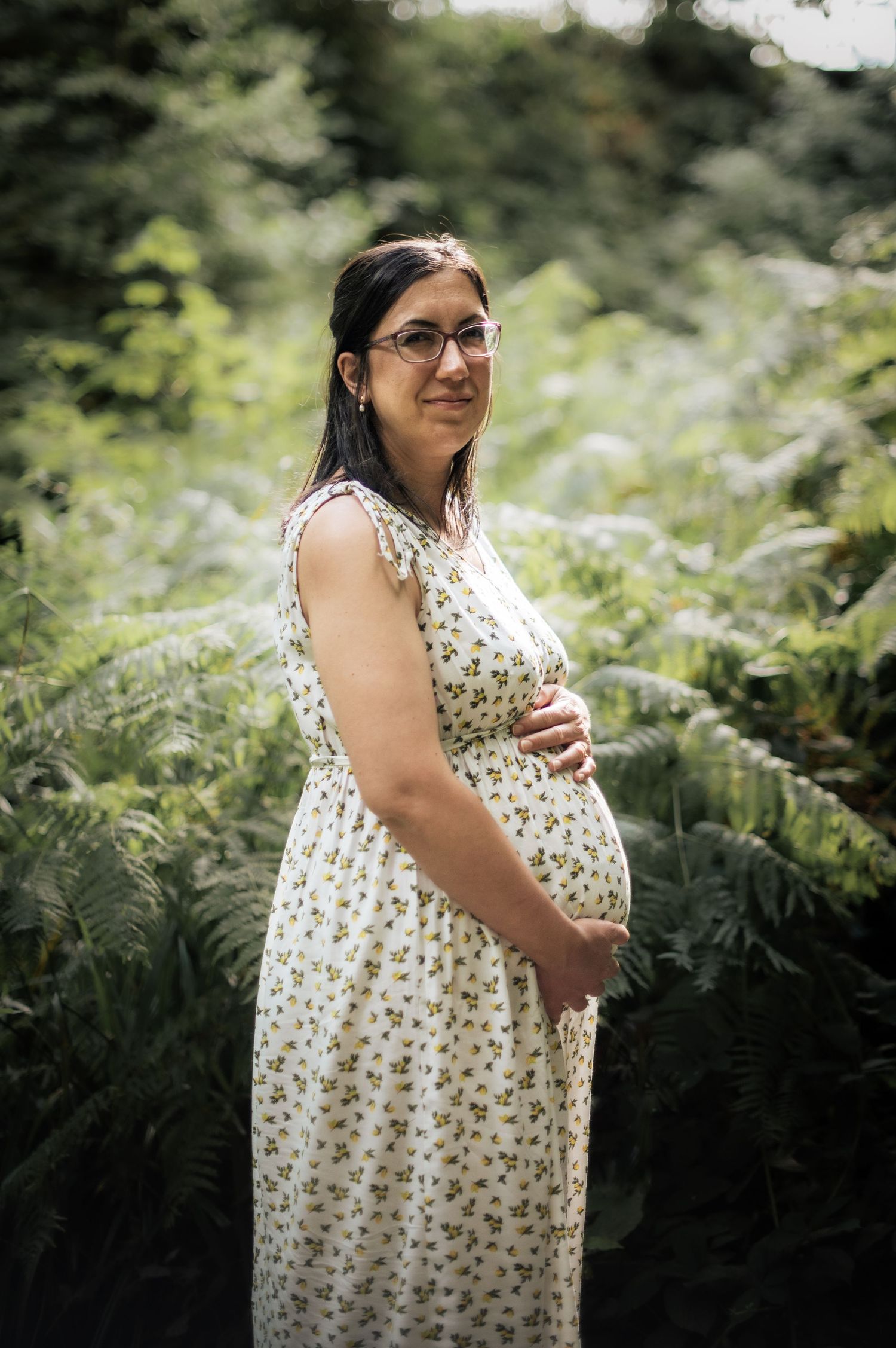 Expectant mother in a floral dress stands among lush green ferns in a park setting during a pregnancy shoot.