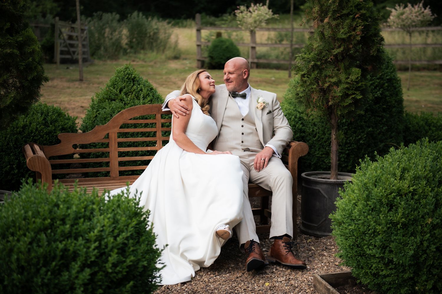 A bride and groom share a romantic moment on a wooden garden bench surrounded by lush green topiary bushes.