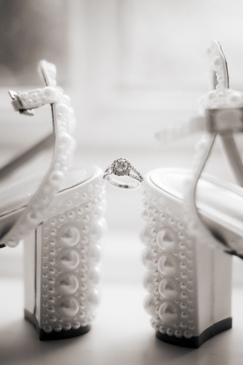 Black and white photo of a diamond engagement ring displayed between pearl-embellished bridal heels.