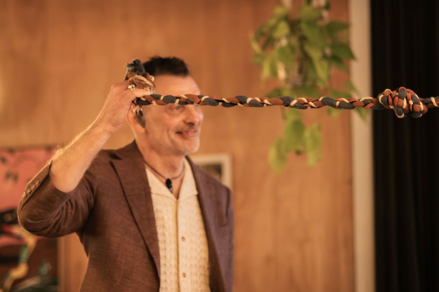 A man in a brown blazer holds a long twisted rope or snake aloft during a magic performance indoors.