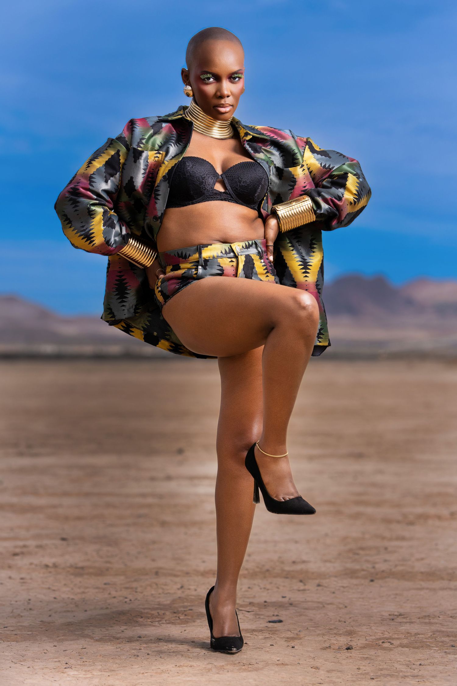 Model poses in colorful patterned jacket and black lingerie against desert landscape with blue sky backdrop.
