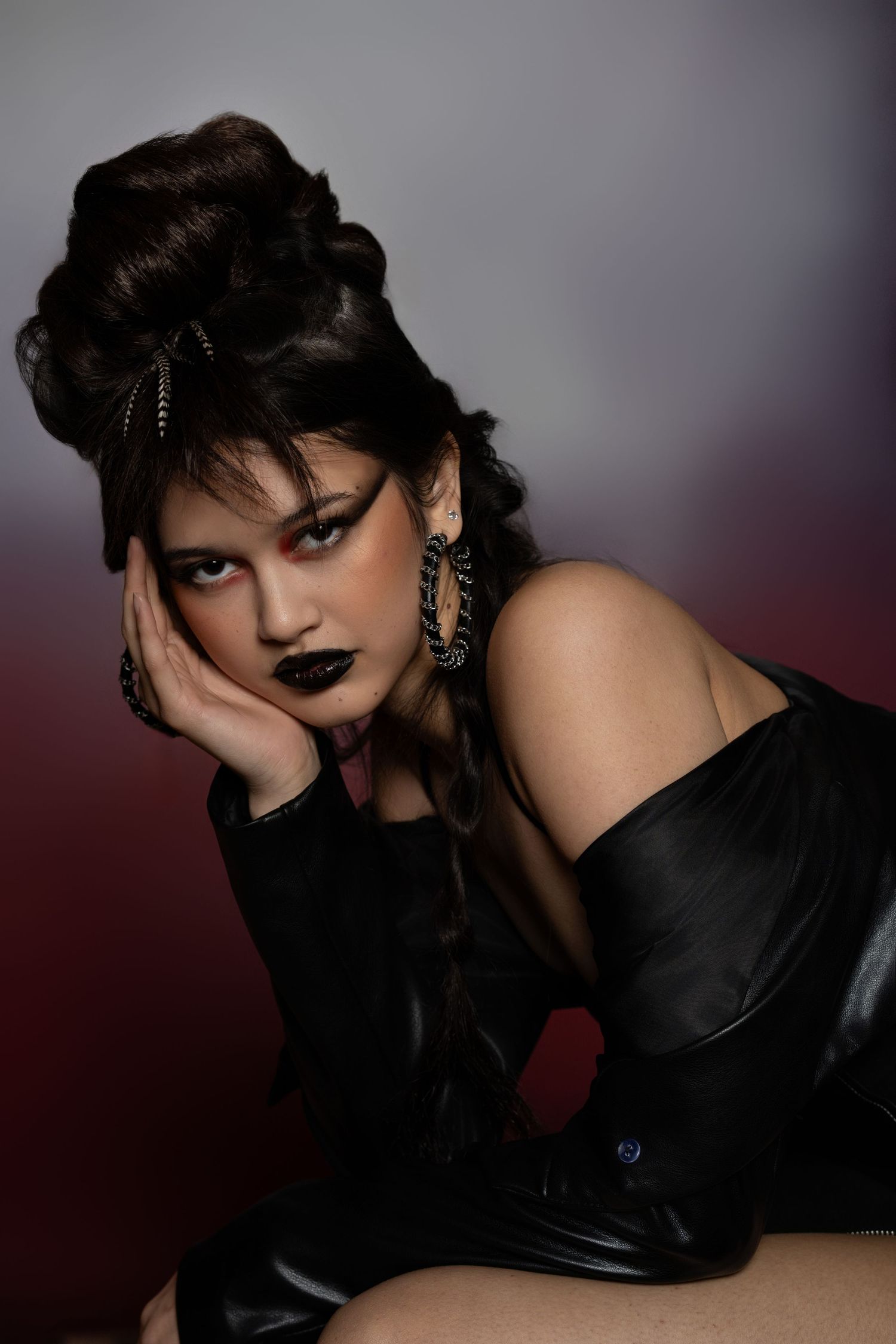 A moody fashion portrait in dark tones features a dramatic updo hairstyle, black gown and gothic makeup against a shadowy backdrop.