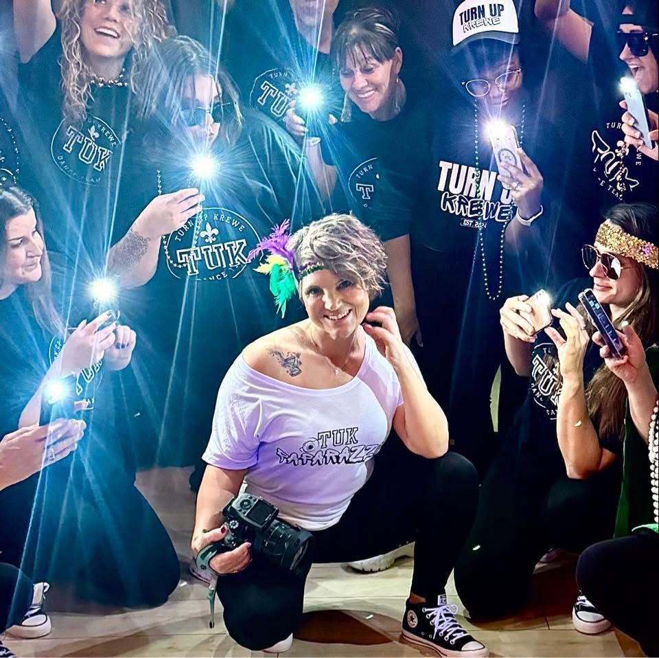 A performer kneeling and posing at an event while fans take photos with phone flashlights in the background.