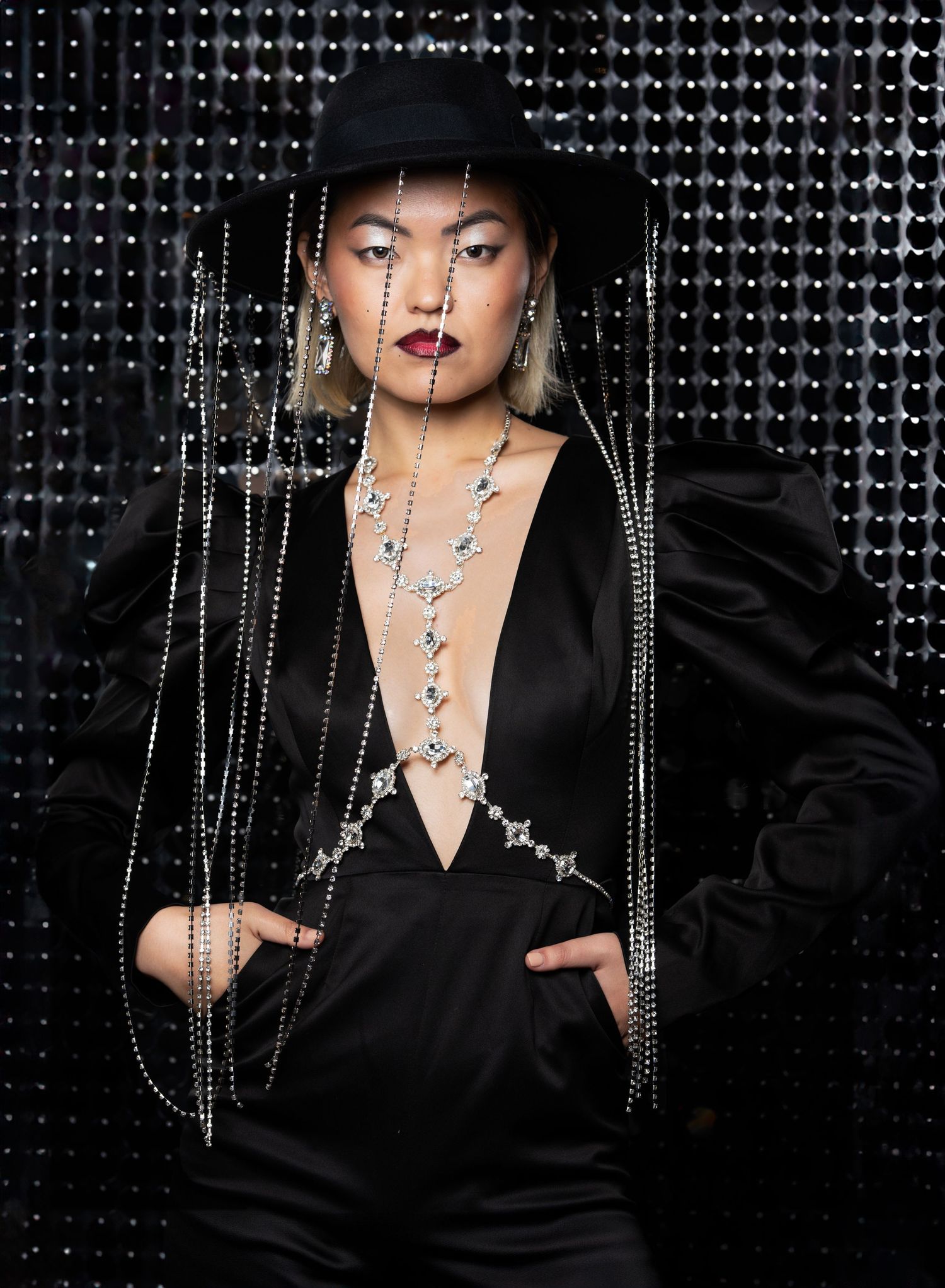 Fashion model in black outfit with dramatic jewelry and hat poses against textured dark background with sparkly chains.