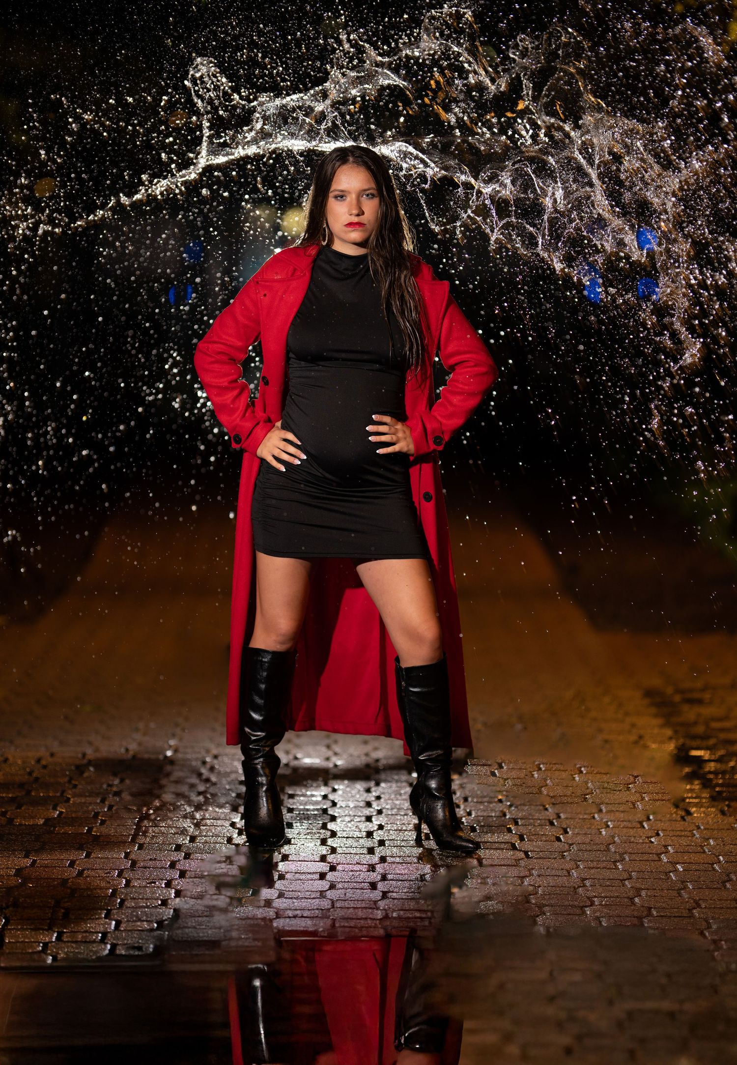 Figure in black dress and dramatic red coat stands on wet pavement with water splash effect and dramatic lighting.