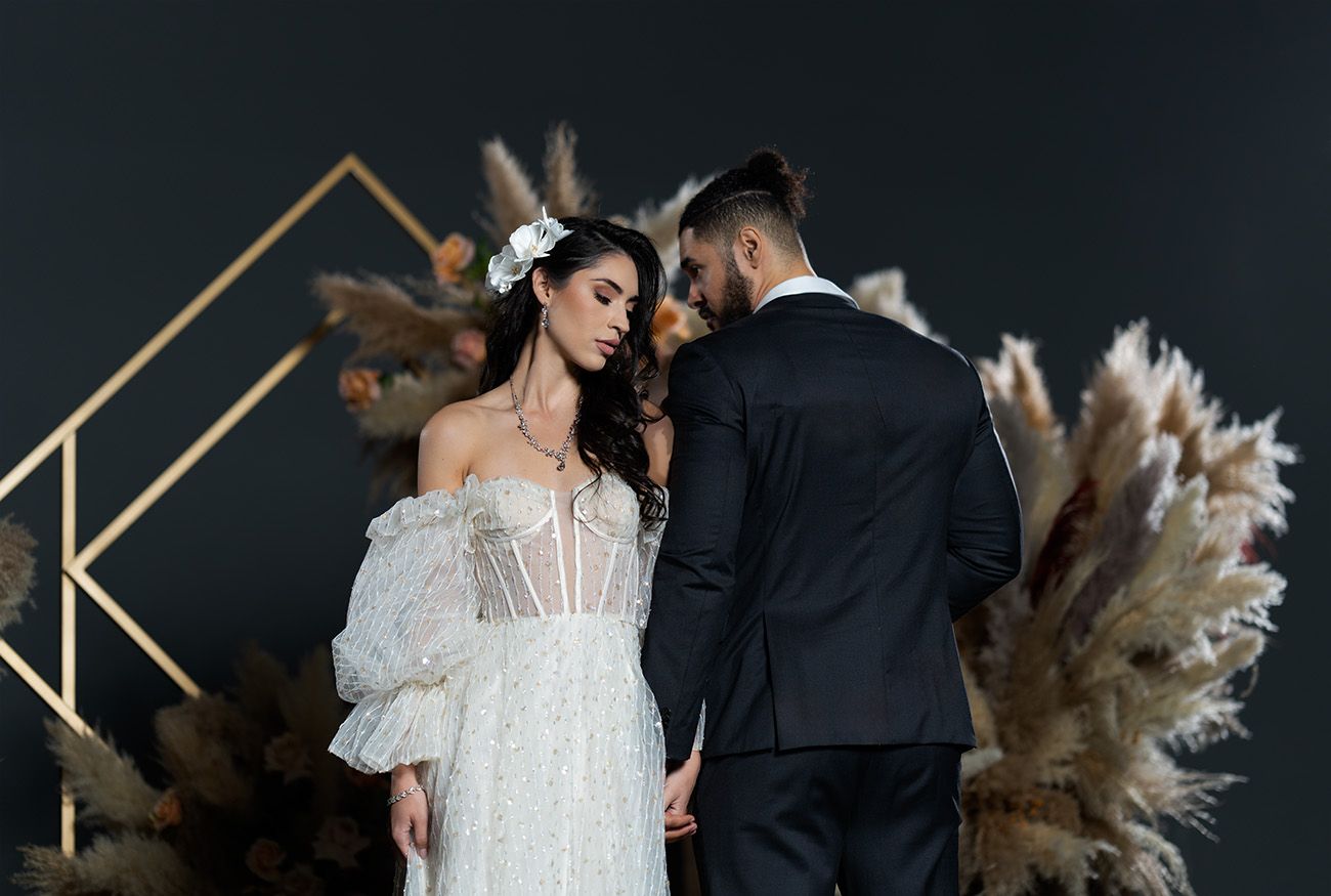 Wedding couple poses against dark backdrop with geometric gold frame and dried pampas grass floral arrangement.