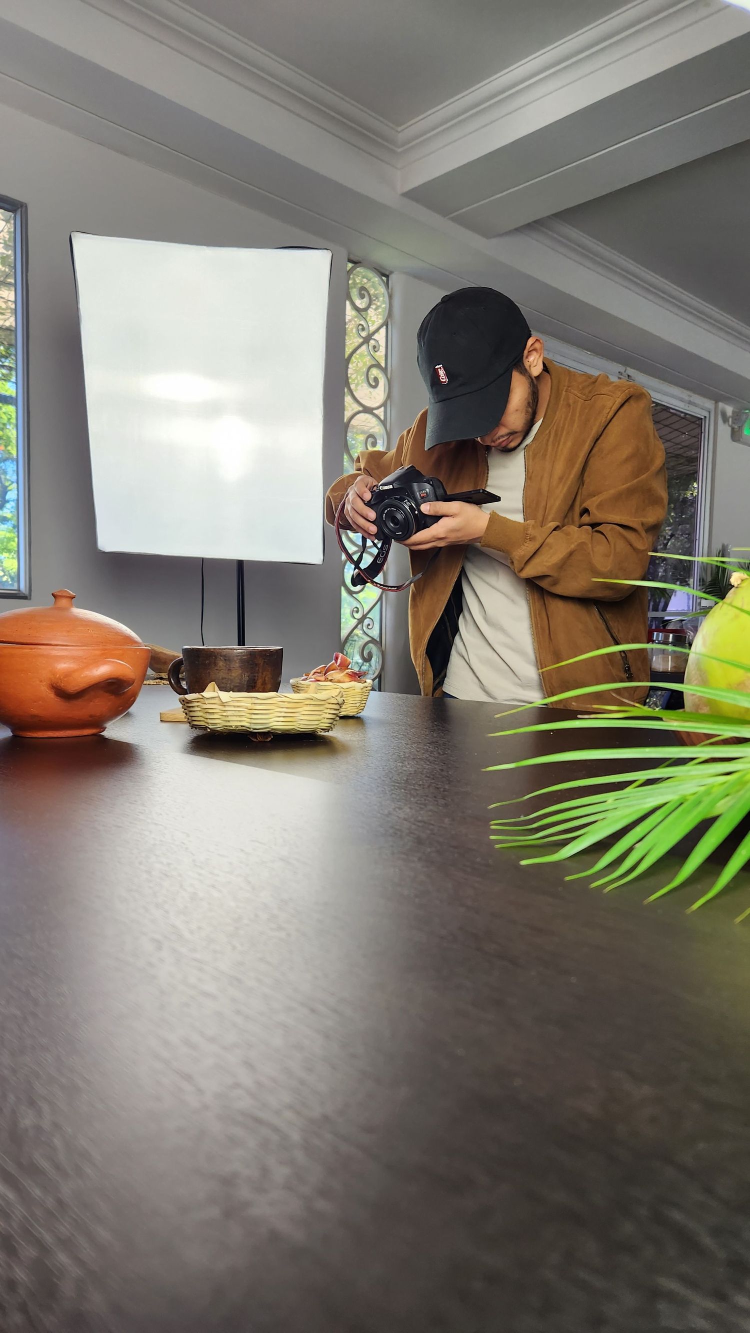 Person in brown jacket and cap photographing food setup on dark table.