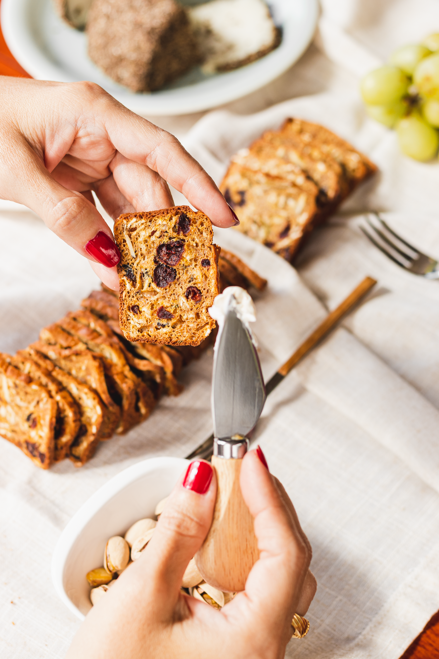 Hands with red nails slice a freshly baked fruit and nut loaf on a white tablecloth.