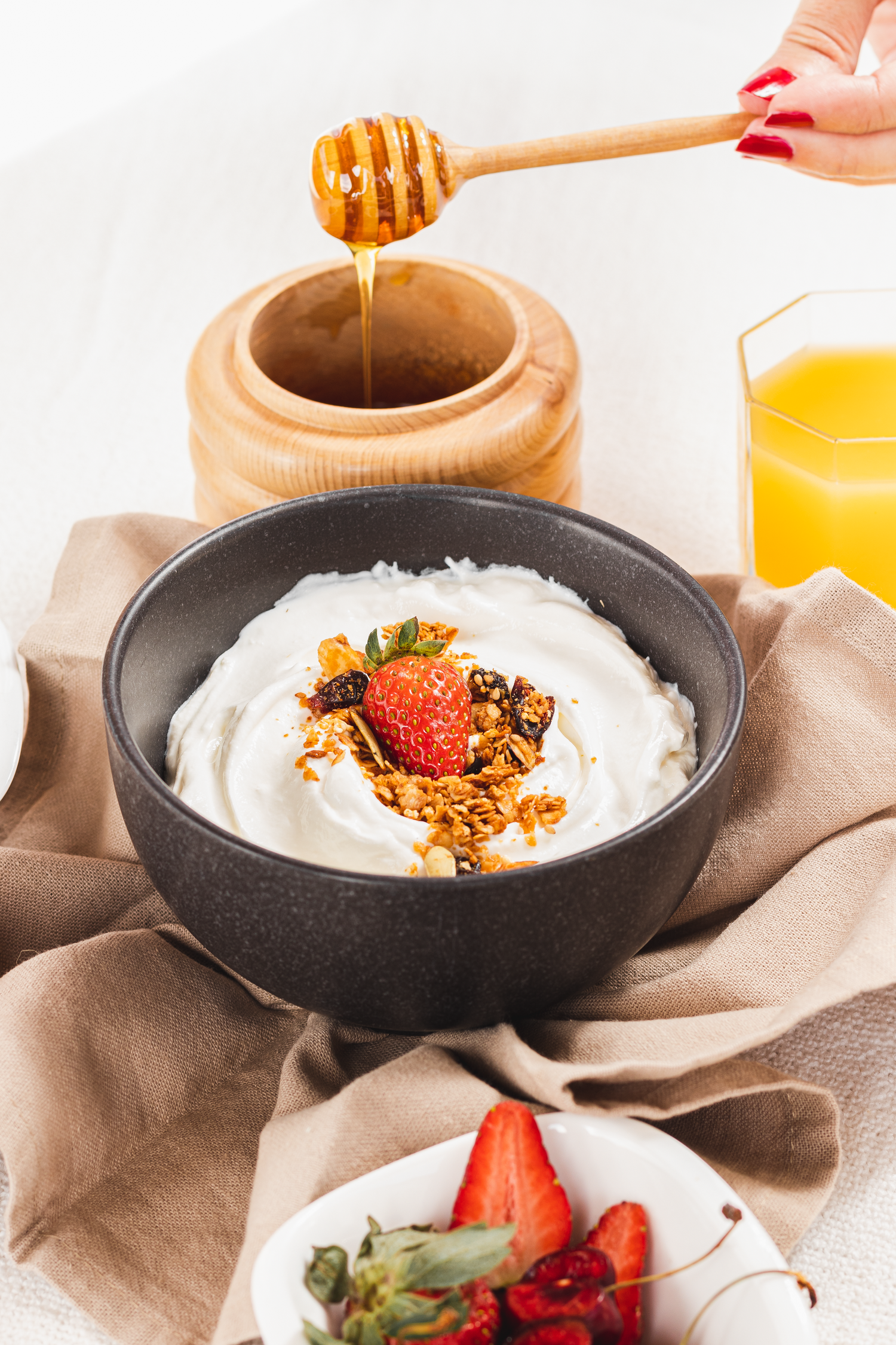 Bowl of yogurt topped with granola and strawberry with honey being drizzled on top.