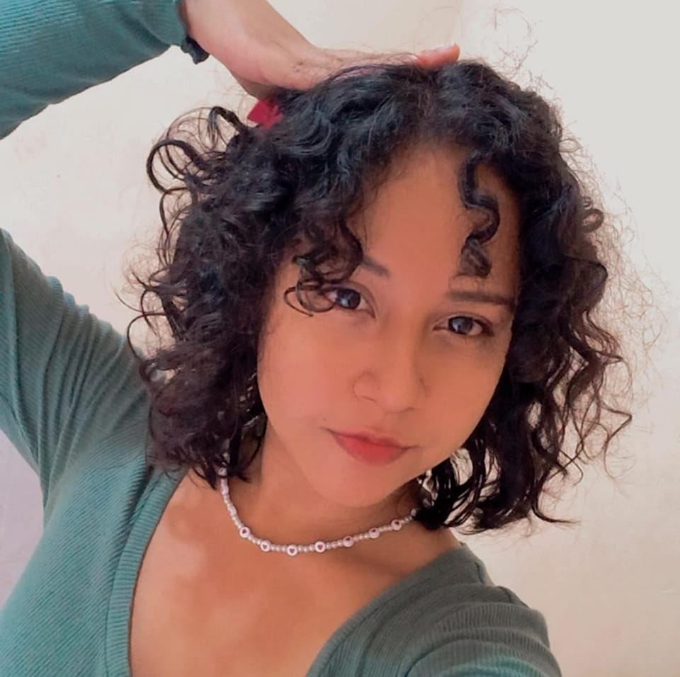 Selfie showing curly black hair, natural makeup, and silver necklace against light background.
