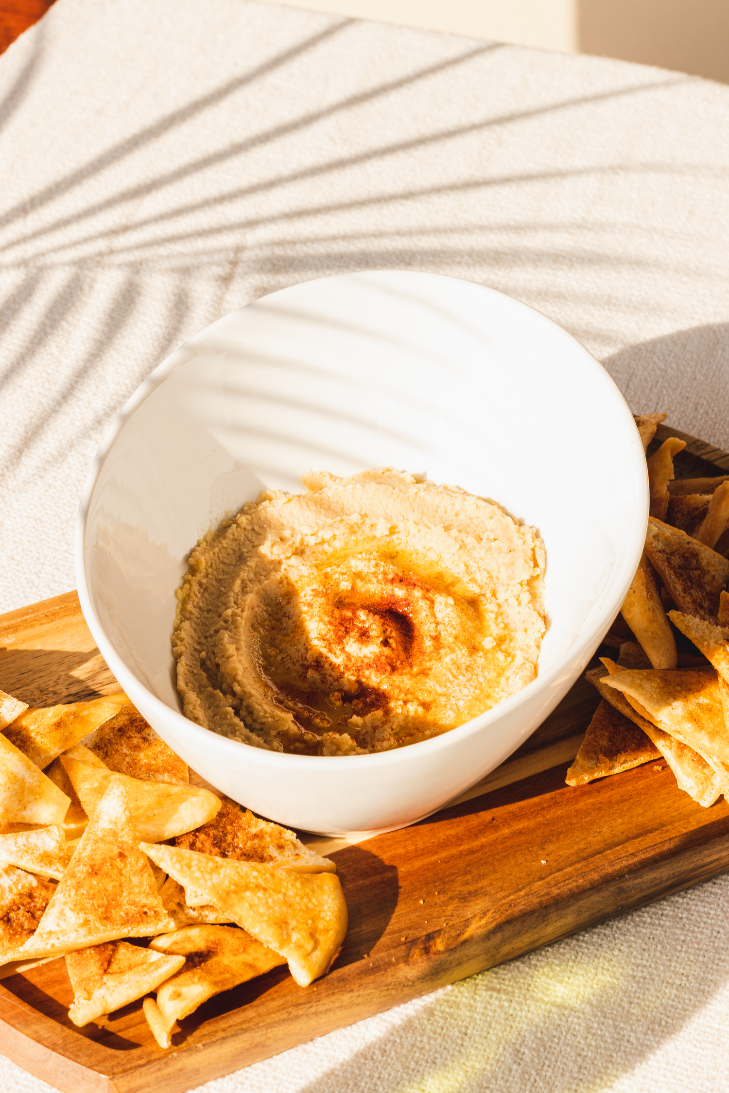 Bowl of creamy hummus with pita chips served on a wooden board in sunlit setting.