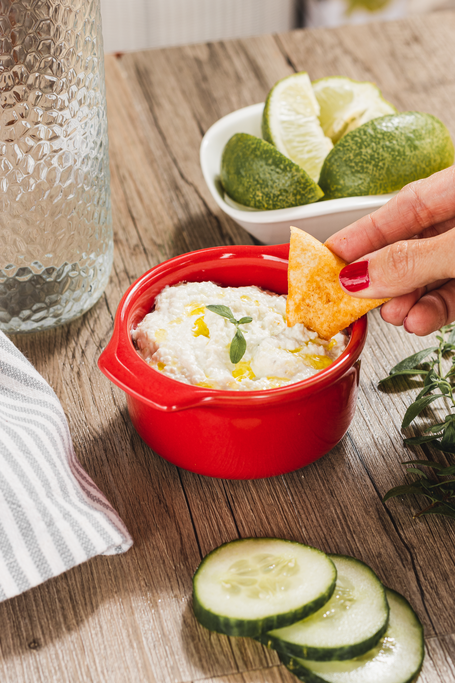 White dip served in red bowl with tortilla chip, limes and cucumber slices on wooden surface.